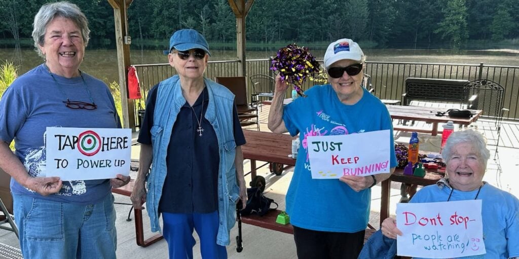 Four women in their 80s and 90s sit and stand outside near picnic tables under an awning and hold signs with cheer to encourage runners in a 5k race. The signs say "tap here to power up" "just keep running" and "don't stop - people are watching"