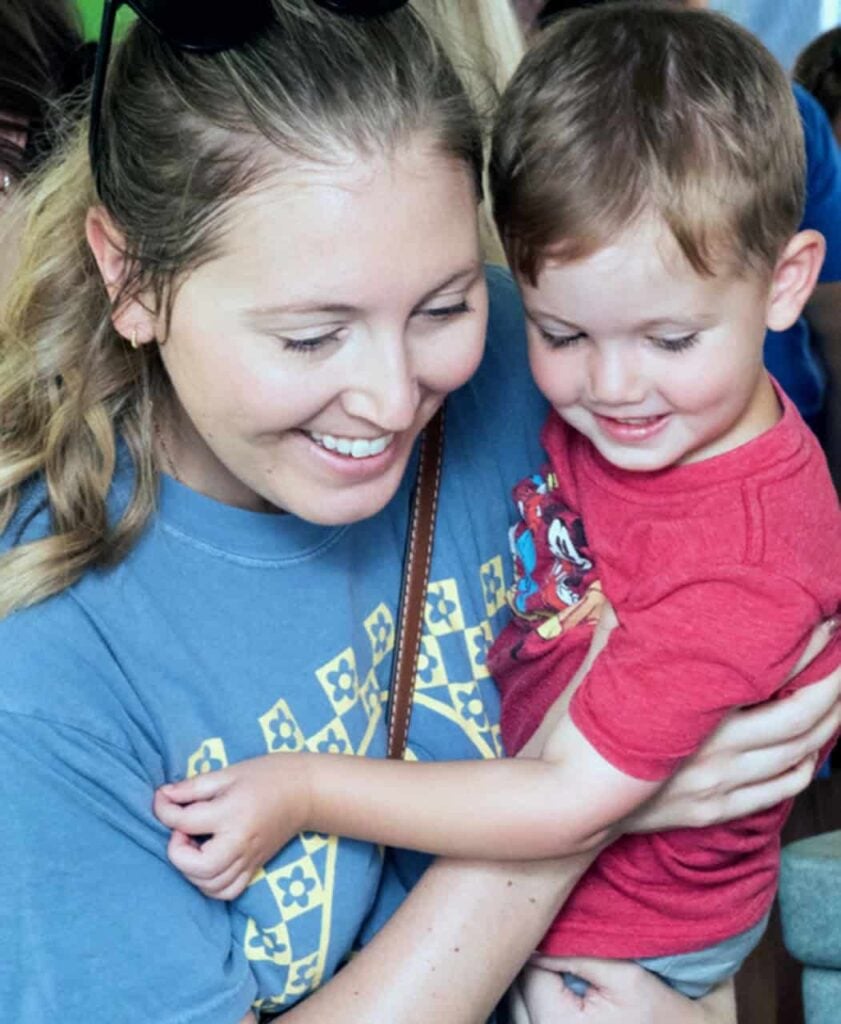 A mom with her hair pulled back in a ponytail wearing a blue shirt holds her small son in a red shirt as they are enthralled looking at animals together at an event.