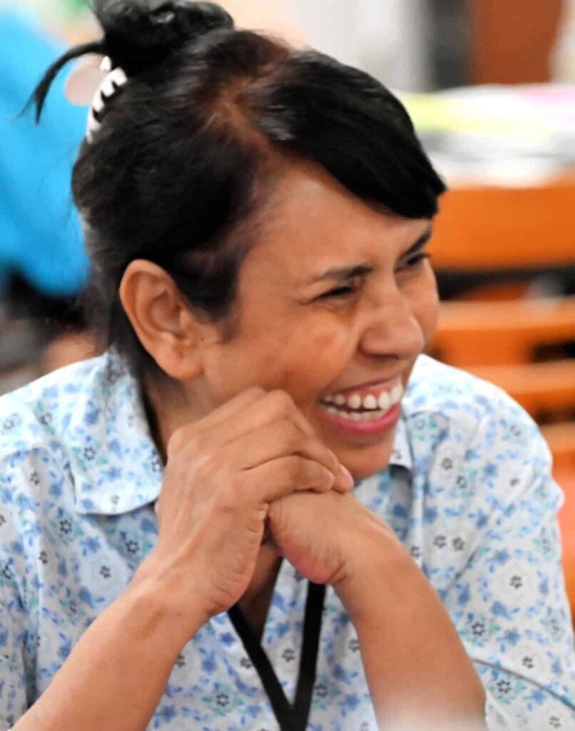 A Pakistani woman with brown hair held up with a white claw clip, wearing a blue flowered shirt is shown laughing at a joke.