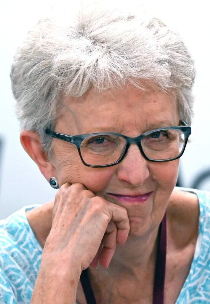 An older white woman with short white hair, blue framed glasses, a blue shirt smiling softly at the photographer as she holds her head up with her hand.