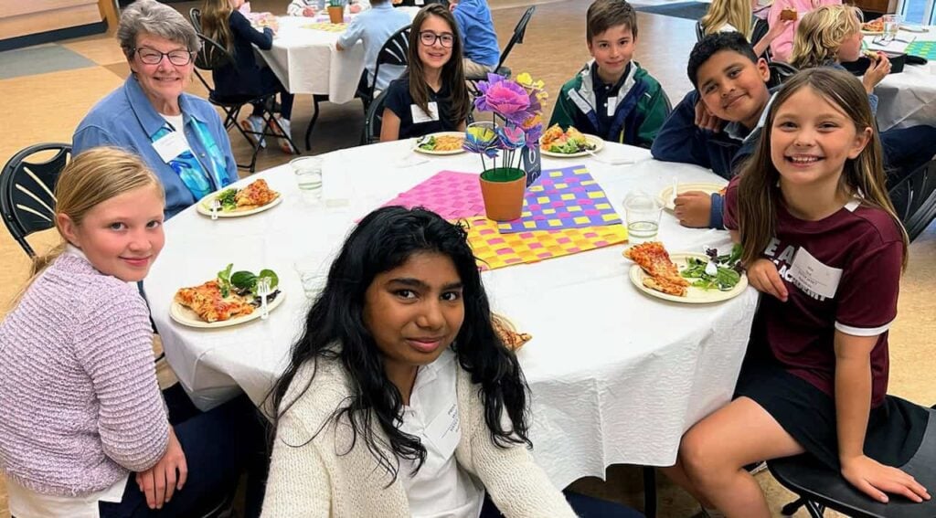 A religious sister with grey hair, glasses and a blue shirt sits with six elementary aged students called "Loretto Leaders" at lunch.