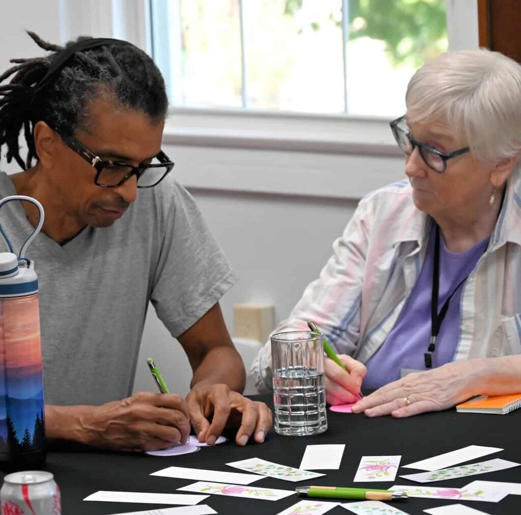 An elder white woman and middle aged Black man work on an activity at a table together.