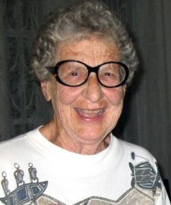 A photo of an elder woman smiling with grey curly short hair, big black oval glasses and a most white t-shirt with an indigenous design on it.
