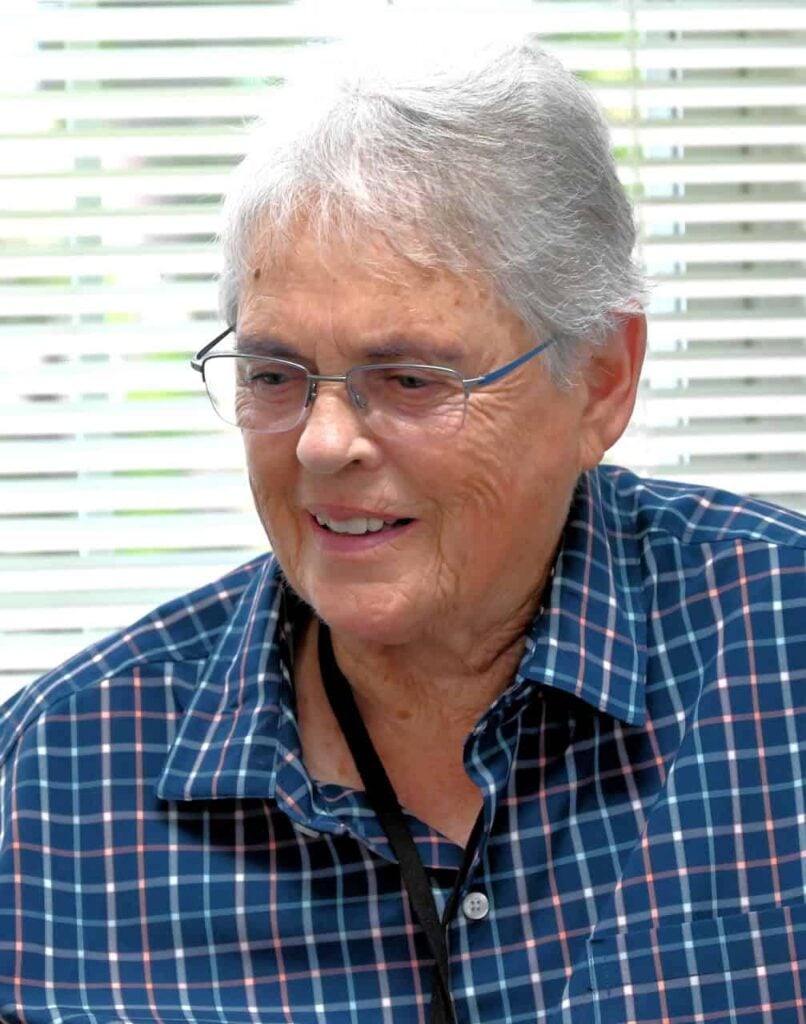 An older white woman with white hair, glasses and a checkered blue shirt ponders during a discussion.