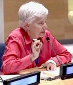 A middle aged woman with short white hair and a pink collard shirt speaking emphatically at the United Nations over a mic.