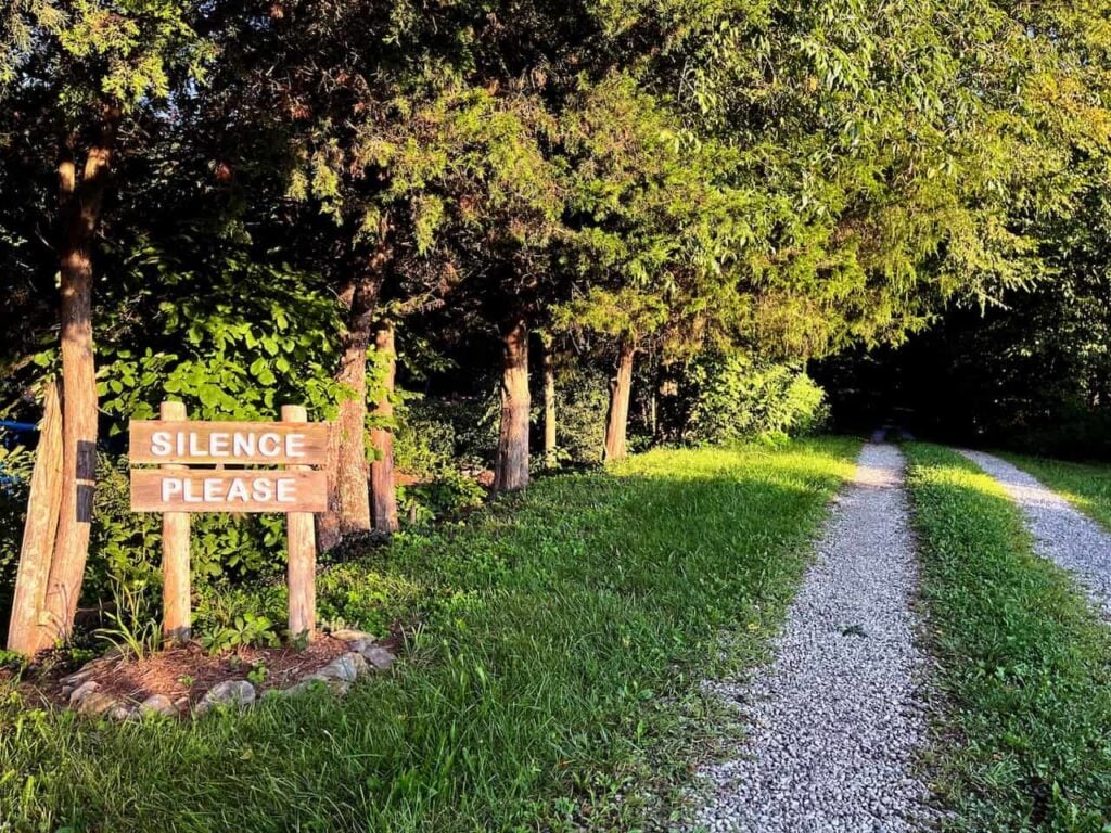 A wooden sign with white text that says "silence please" at the beginning of a gravel road leading into the woods full of thick green trees.
