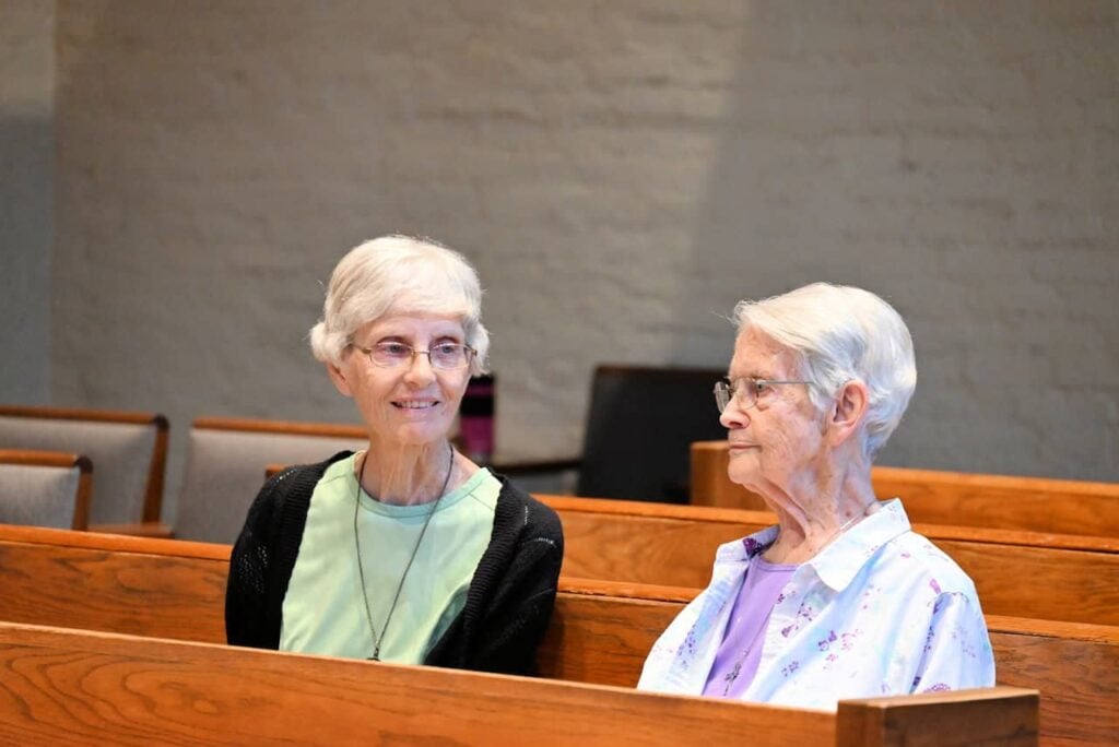 Two women in their 80s sit in a wooden church pew talking. They have short white hair and glasses. One woman on the left is wearing a green shirt and black cardigan. The woman on the right is wearing a purple shirt with a light purple flowered shirt over it.
