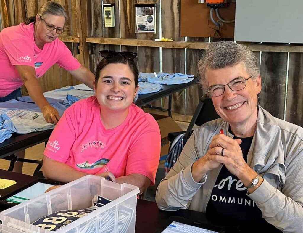 Two women smiling sitting at a table and one woman behind them laying out blue t-shirts on another table. They are ready to help 5k participants collect their numbers and t-shirts for a race.