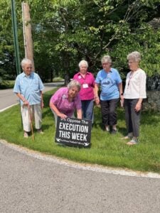 Five women in their 80s and 90s standing in the grass as one woman bends down and puts a black sign with white text that says "We oppose the execution this week forwhomethebellstoll.org"