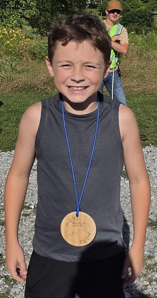 An elementary school aged kid with a grey tank top and black shorts smiles after winning a 5k in his age category. He also wears a wooden medal on a blue string around his neck.