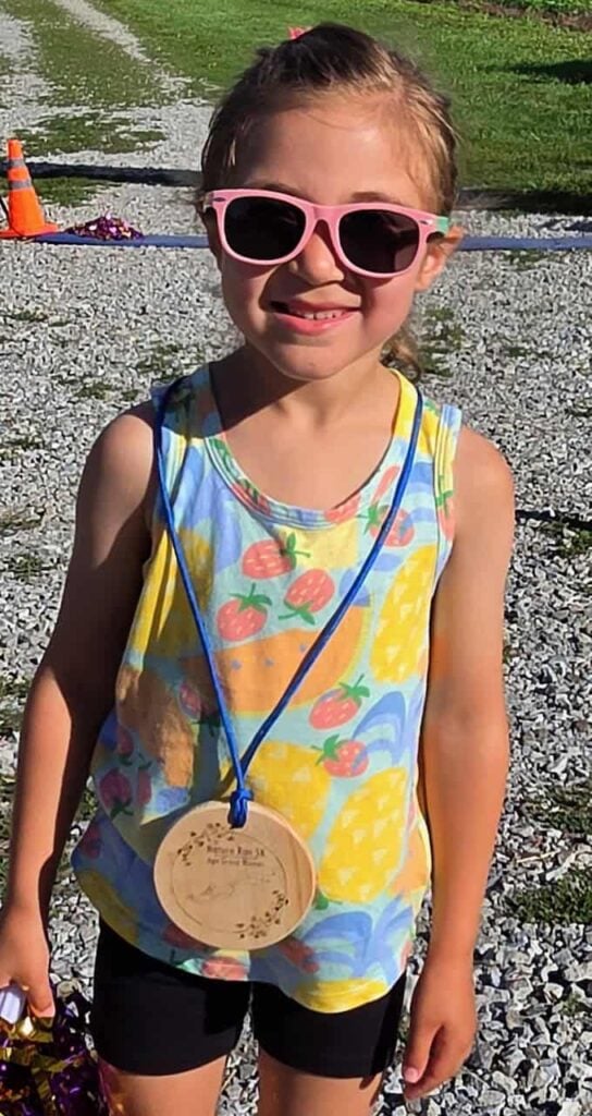 An elementary school aged kid with pink sunglassess, a shirt with fruit on it and black shorts smiles after winning a 5k in her age category. She also wears a wooden medal on a blue string around her neck.