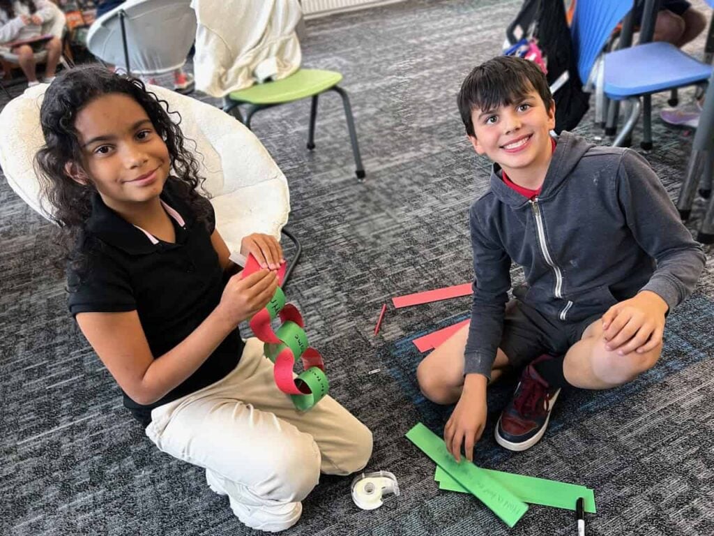 Two elementary aged students, a girl and a boy smile mid craft. They are making paper chains out of green and red paper that have writing on them.