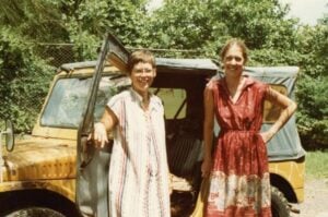 Two women in light Chilean cotton dresses pose in front of a yellow Jeep on a dirt road surrounded by trees.