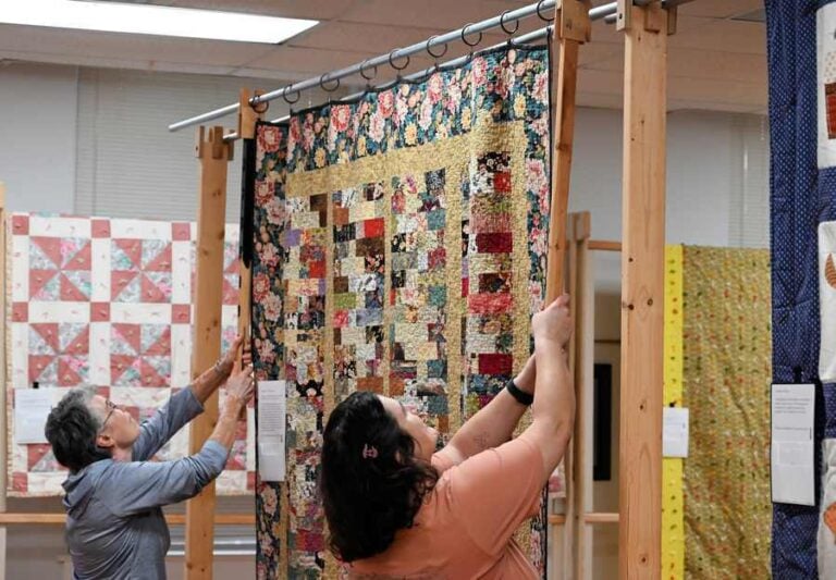 Two women use long wooden tools with cutouts at the top to lift from a rack the pole on which a large, colorful patchwork quilt is hanging for display.