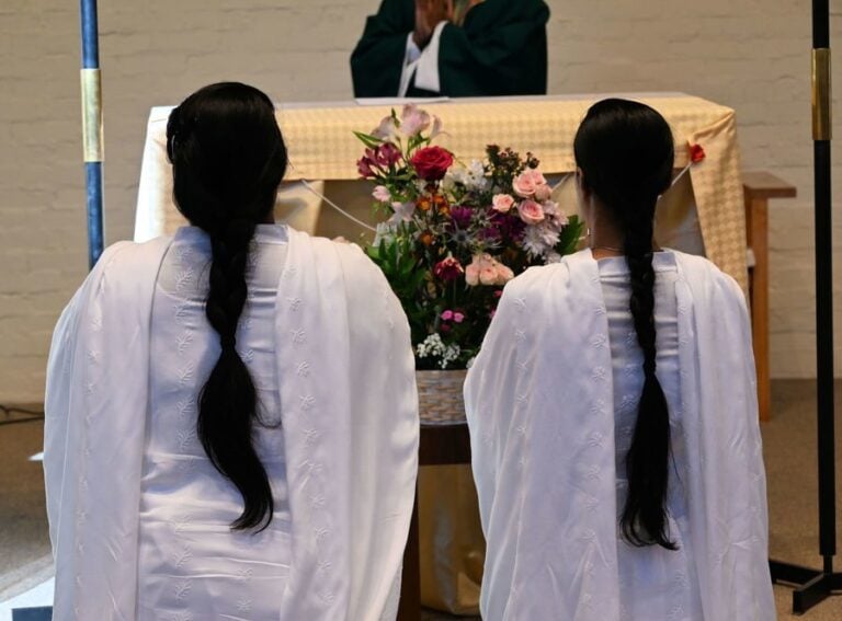 Two women with long dark braids and wearing long white garments kneel before a flower-bedecked altar. A priest is partially visible standing behind the altar.