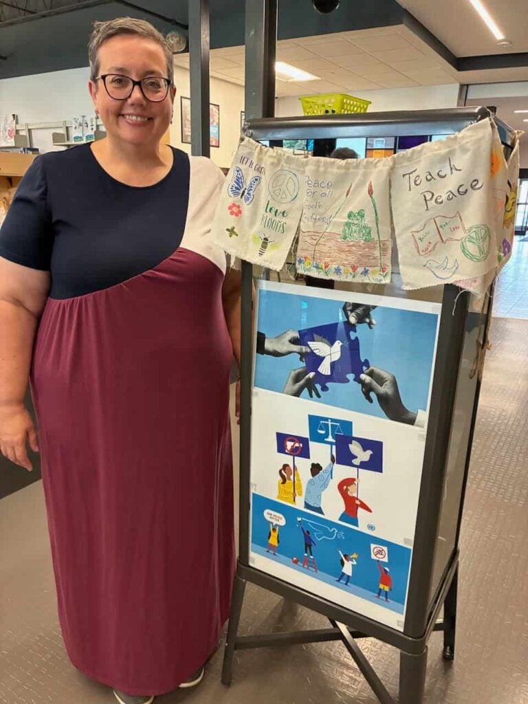 A woman with short hair and glasses and a blue, white and red dress standing next to a display of peace at the library for International Peace Week.