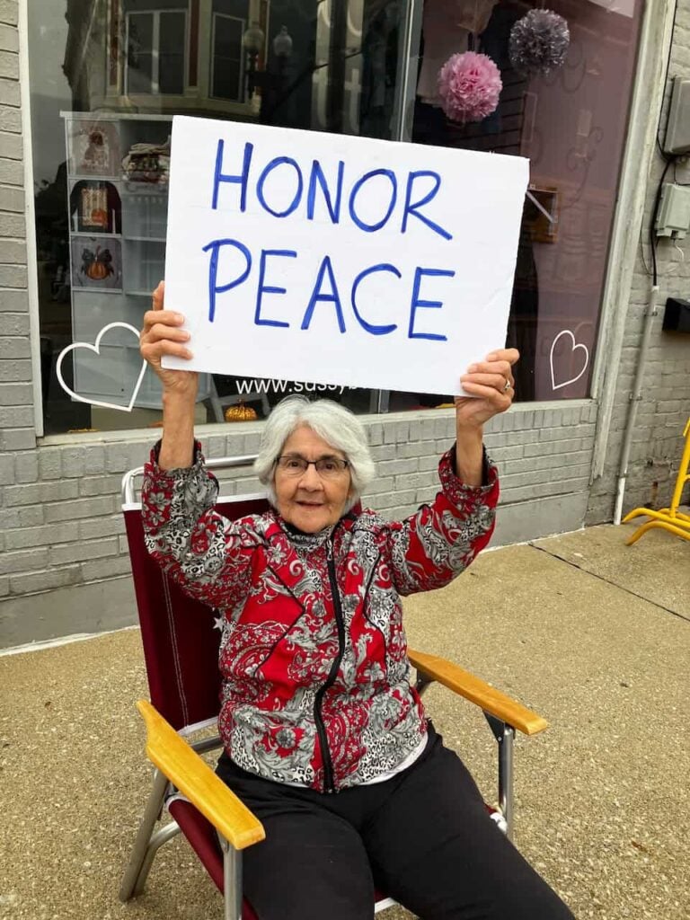 An elder woman with white hair and a colorful red and grey jacked and black pants sits on a folding chair on a street corner and holds up a sign that says "Honor Peace"