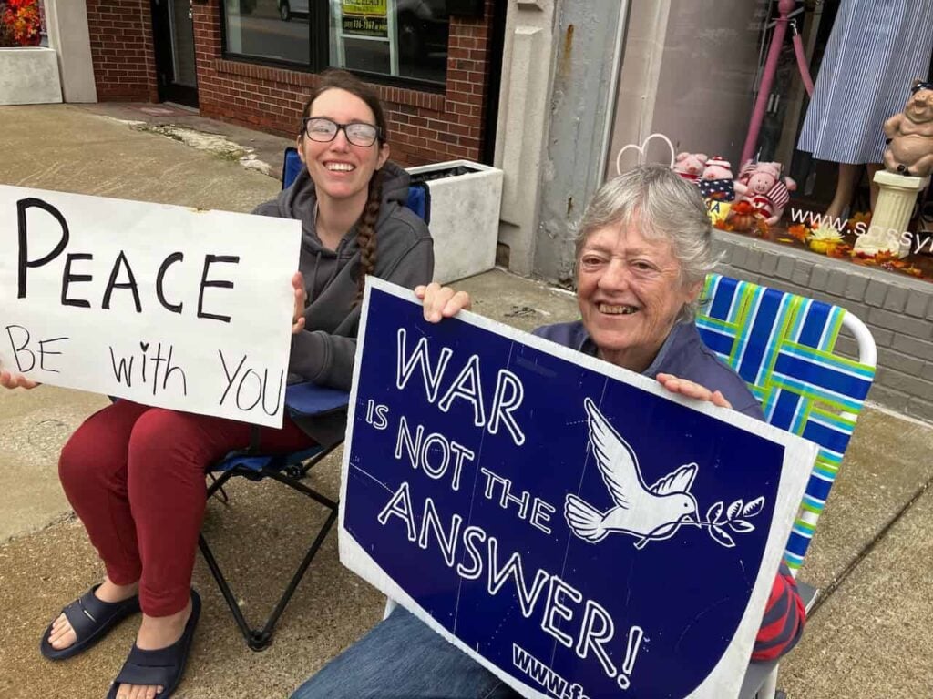An elder nun sitting in a lawn chair on the corner of an intersection sits next to a younger woman and they are both holding signs of peace. One says "Peace be with you" the other says "War is not the answer"