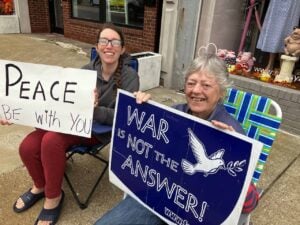 An elder nun sitting in a lawn chair on the corner of an intersection sits next to a younger woman and they are both holding signs of peace. One says "Peace be with you" the other says "War is not the answer"