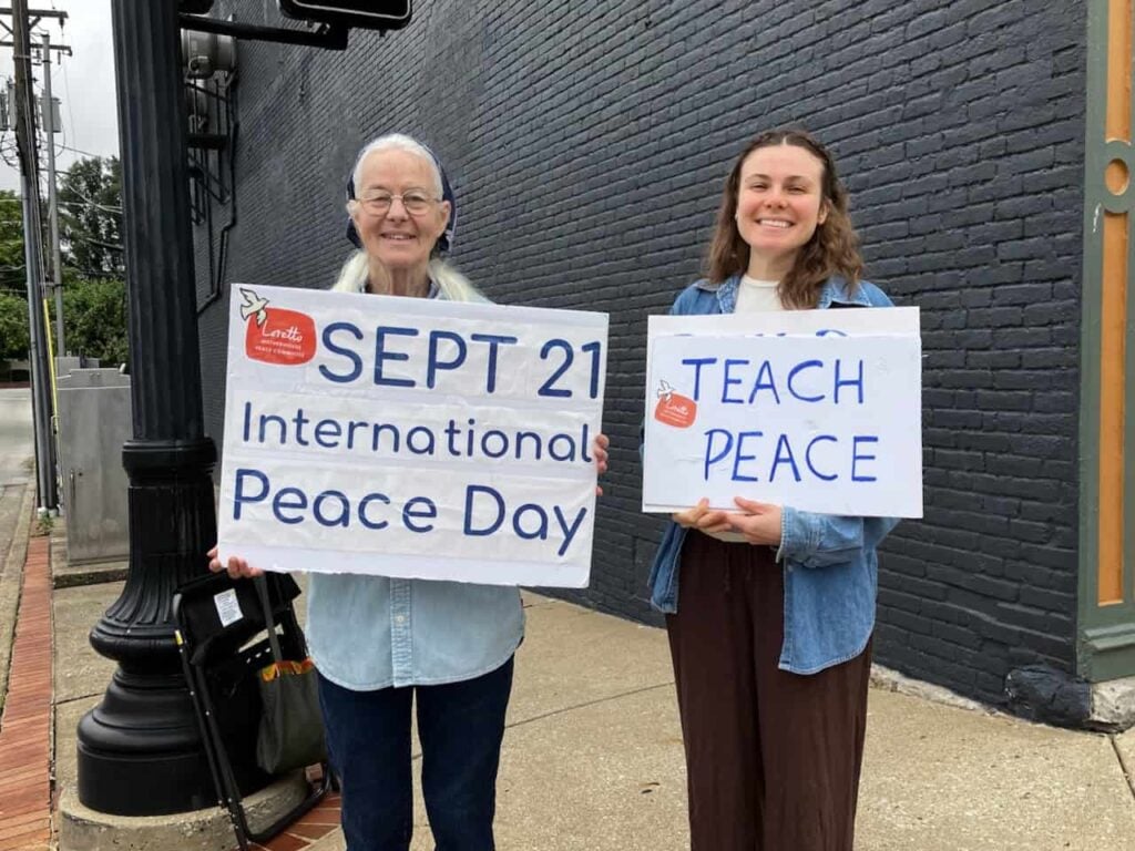 An older woman next to a younger woman standing smiling on a street holding signs about peace. One sign says "Sept. 21 international peace day" and the other says "teach peace"