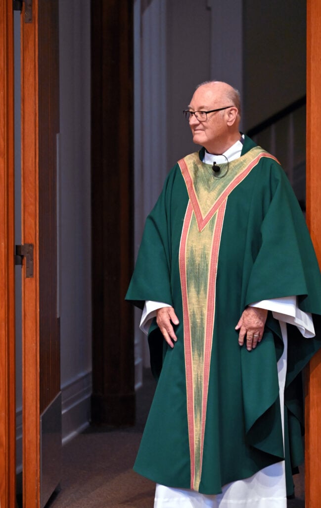 A priest in green and gold vestments stands in a church door to welcome Mass attendees