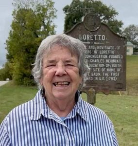 An older woman smiles for the camera while standing outside next to a sign titled "Loretto" followed by details of the Sisters of Loretto history.