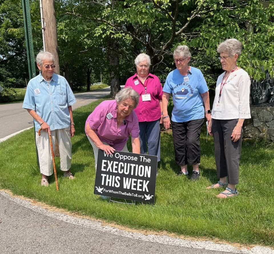 Five women in their 80s and 90s standing in the grass as one woman bends down and puts a black sign with white text that says "We oppose the execution this week forwhomethebellstoll.org"