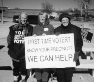 A black and white photo of three older women dressed in very warm clothes standing outside smiling holding a sign that says "First time voter? Know your precinct? We can help"