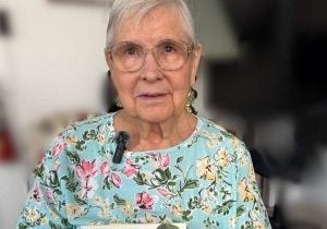 An older woman wearing a flowered shirt with a microphone attached to the collar holds up a book by Joyce Rupp.