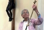 An older religious sister with short grey hair, glasses, a white shirt and pants and a purple patterned cardigan holding a rope to toll a church bell.