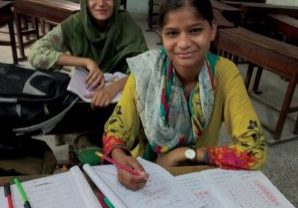 Photo of two Pakistani middle aged girls wearing colorful clothing, one in green patterns and the other with yellow, red, green and black patterns smiling as they take a break from working on their school work which is displayed on the desk in front of them.