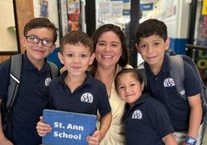 Four elementary students in school uniform polo shirts and big smiles gather round Kathleen Fox, principal at St. Ann's School, for a group photo.