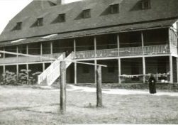 Archival photo of large two story building with a balcony running the entire length of the second floor. A habited nun is standing in front of the building, next to a clothesline.