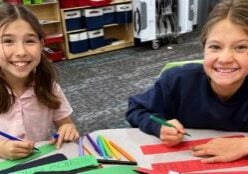Two elementary aged girls take a break to smile for a picture as they are holding markers andd making placemats and paper floral centerpieces for an upcoming event.