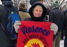 A woman in a heavy black jacket and wool gloves stands in a crowd with snow on the ground holding a red banner that says "violence has never defeated violence"