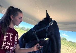 Young woman with long ponytail smiles looking down at a horse with the halter in her hands.