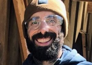 A white man with a dark beard smiling outside as he wears clear working goggles, a tan beanie and blue sweatshirt.