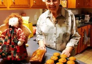 A smiling woman stands at her kitchen counter with a rustic angel doll next to cooling rack with a loaf of bread and muffins.