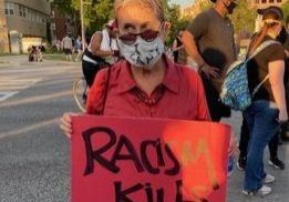 Woman in sunglasses and mask stands in the street holding a red sign that reads "Racism Kills."