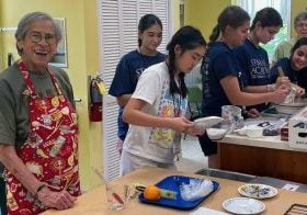 A couple older women are pictured helping high school students make scones in a kitchen.