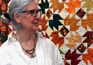 An older white woman with grey hair in a bun and a white shirt and turquoise jewelry with black glasses stands in front of a orange pattered quilt.
