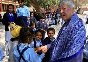 A short-haired woman in a blue shall stands holding hands with young school children outside on a sunny day.