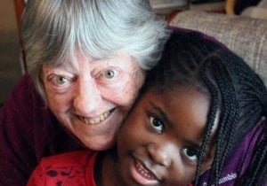 An older white woman with grey hair smiles with cheeks pressed together with a young Black girl with long cornrows in two pigtails.