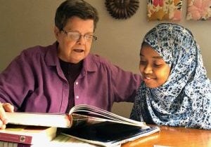 An older white woman with brown hair and glasses and a pink shirt is shown helping a young woman of color with her homework. She is wearing a beautiful blue and white patterned hijab.