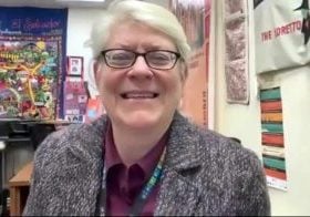 A middle-aged woman with short white hair and small rectangular glasses is smiling as she sits in an office decorated with bright colorful posters behind her.