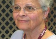 A woman with metal framed glasses and white hair wearing stud silver earrings giving a soft smile not looking at the camera. She is wearing a blue shirt with a lanyard that has the word "Loretto" repeating.