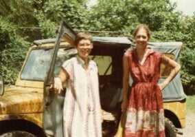 Two women in light Chilean cotton dresses pose in front of a yellow Jeep on a dirt road surrounded by trees.