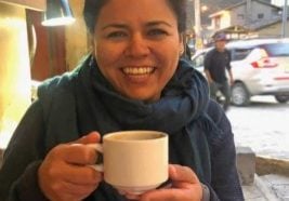 A Latina woman in her late 30ss early 40s smiles holding a cup of coffee outside a coffee shop. She is wearing a blue sweater and scarf and her black hair is in a high ponytail.