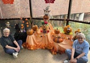 Two gray-haired women sit on the floor flanking a festive Halloween display of fall colors, foliage and pumpkins with sweet, painted jack o' lantern faces.