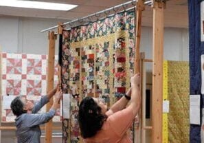 Two women use long wooden tools with cutouts at the top to lift from a rack the pole on which a large, colorful patchwork quilt is hanging for display.